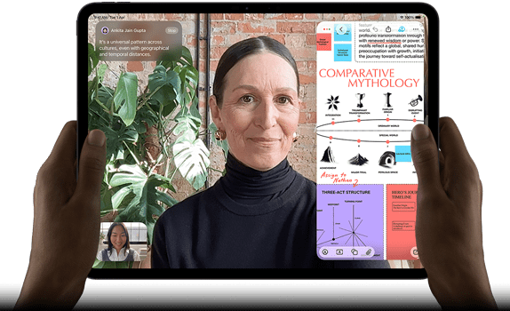 iPad Pro, front exterior, landscape orientation, space black color, rounded corners, black display bezel, hands holding left and right sides, display showing a FaceTime conversation being live translated, and an educational app showing a presentation of "Comparative Mythology" with text, diagrams, and handwritten notes