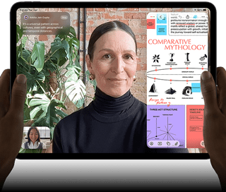 iPad Pro, front exterior, landscape orientation, space black color, rounded corners, black display bezel, hands holding left and right sides, display showing a FaceTime conversation being live translated, and an educational app showing a presentation of "Comparative Mythology" with text, diagrams, and handwritten notes